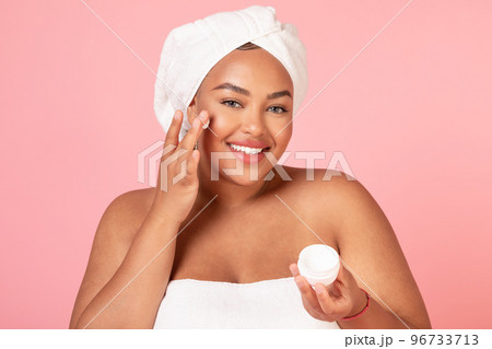 Beautiful african american body positive lady holding jar of cream, and applying moisturizer on cheek, pink background 96733713