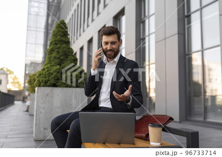 Smiling busy young european man manager with beard in suit with cup of coffee and laptop calls by phone 96733714
