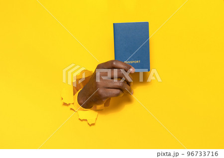 African american traveler tourist male hand holding passport from a torn hole in yellow paper wall background African american traveler tourist male hand holding passport from a torn hole in yellow paper wall background 96733716