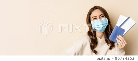 Travel and covid-19 pandemic. Close up portrait of smiling woman in medical mask, showing passport with two tickets, concept of tourism, beige background Travel and covid-19 pandemic. Close up portrait of smiling woman in medical mask, showing passport with two tickets, concept of tourism, beige background 96734252