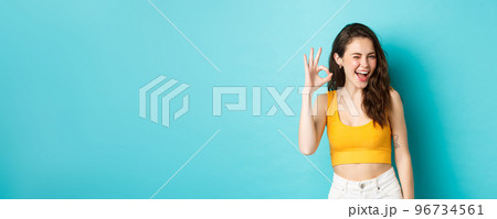 Young cheerful lady in summer clothes winking and saying yes, alright gesture, making OK sign in approval, standing against blue background 96734561