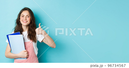 Young woman attend courses, girl student studying, holding notebooks and showing thumb up in approval, recommending company, standing over blue background 96734567