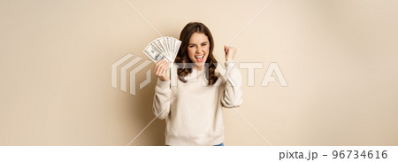 Enthusiastic modern woman winning money, got cash, celebrating and shouting of joy, standing against beige background 96734616