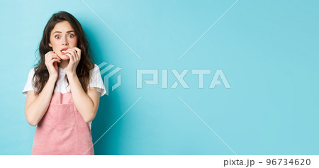 Portrait of scared and startled brunette girl biting fingernails, staring at camera shocked and worried, hear terrible news, standing in stupor against blue background Portrait of scared and startled brunette girl biting fingernails, staring at camera shocked and worried, hear terrible news, standing in stupor against blue background 96734620