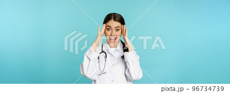 Portrait of surprised and excited smiling doctor woman, holding hands on head, reaction to mindblowing awesome news, standing over blue background Portrait of surprised and excited smiling doctor woman, holding hands on head, reaction to mindblowing awesome news, standing over blue background 96734739