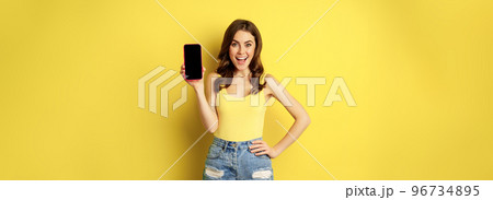 Stylish smiling woman showing mobile phone screen, app interface on smartphone, standing in tank top and shorts against yellow background 96734895