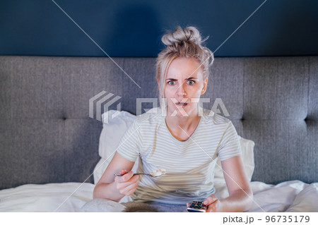 Shocked, stressed, surprised woman eating ice cream, holding a remote control while watching TV in bed. Scary horror movie or terrible news, scare in her eyes Overeating, Jamming of negative emotions. 96735179