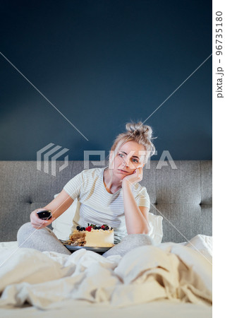 Bored, sad woman in home clothes eating cake and using the remote control to change the channel while watching television in bed at night. Overeating, Jamming of negative emotions. Vertical card. 96735180