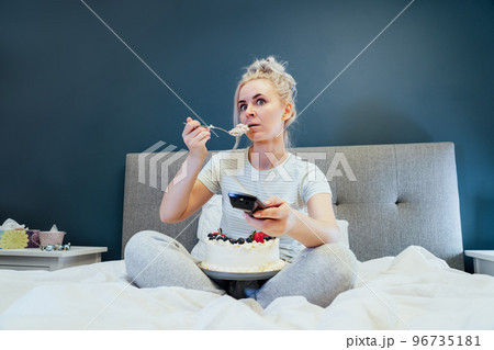 Surprised, stressed, shocked woman eating cake and holding a remote control while watching TV in bed. Scary horror movie or terrible news, scare in her eyes Overeating, Jamming of negative emotions 96735181
