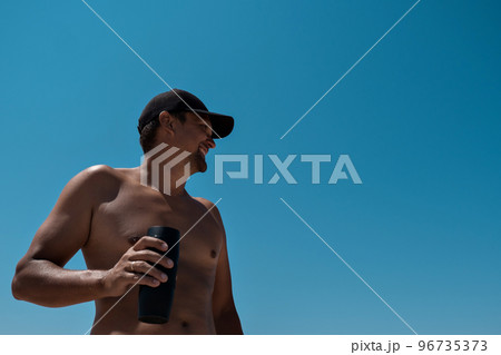 A tanned man in a black cap and with a naked torso drinks coffee on the beach against the blue sky. Recreation enjoyment. Athlete drinks water after a run. High quality photo. 96735373