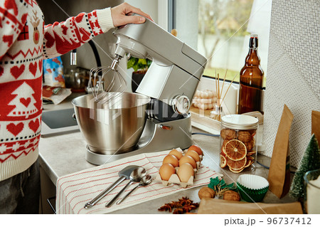 Woman in festive christmas sweater preparing dough for cookies at home kitchen. Female hands use electric mixer. Modern kitchen household appliances Woman in festive christmas sweater preparing dough for cookies at home kitchen. Female hands use electric mixer. Modern kitchen household appliances 96737412