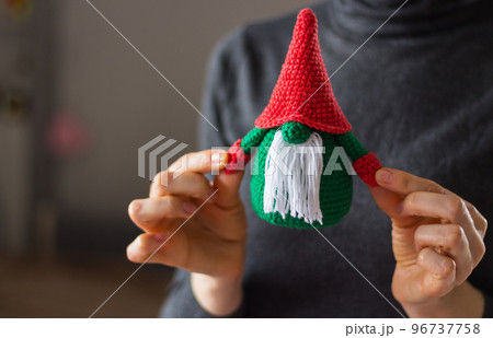 Funny knitted toy in woman hands. Girl holding Christmas handmade gift. Cute dwarf with white beard. Winter decoration. Happy New Year present. Knitted Santa toy in girl's hands. Christmas symbol.  96737758