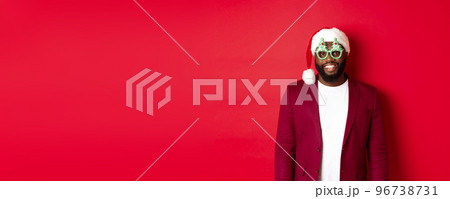 Funny Black man in santa hat and party glasses celebrating Christmas, smiling happy and wishing merry xmas, standing over red background Funny Black man in santa hat and party glasses celebrating Christmas, smiling happy and wishing merry xmas, standing over red background 96738731