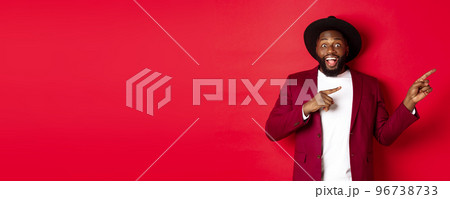 Winter holidays and shopping concept. Stylish african american man pointing fingers right at copy space for logo, standing over red background 96738733