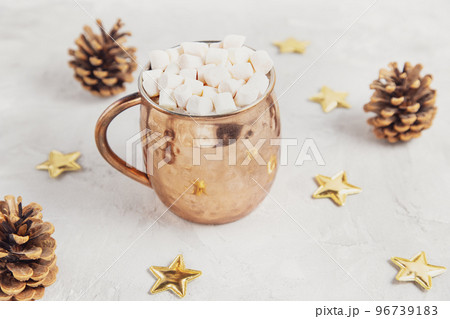 Copper mug with cocoa or chocolate and marshmallows on top, gold stars and pine cones Christmas decoration on light gray background. Winter hot beverage concept, copy space 96739183