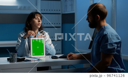 Physician holding tablet computer pointing at green screen chroma key template while discussing patient disease report with assistant. Team working over hours at medical expertise in hospital office 96741126