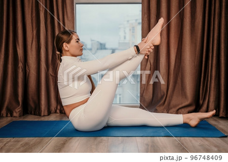 Well looking middle aged woman with long hair, fitness instructor in white sportswear, doing stretching and pilates on yoga mat at home. Healthy lifestyle, harmony, fitness yoga and meditation. 96748409
