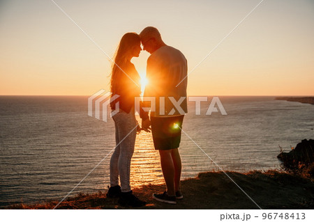 Silhouette of a young romantic couple enjoying an evening on a cliff above the sea with a red burning sunset. Traveling couple, man and woman, holding hands and standing on top of a mountain at sunset Silhouette of a young romantic couple enjoying an evening on a cliff above the sea with a red burning sunset. Traveling couple, man and woman, holding hands and standing on top of a mountain at sunset 96748413