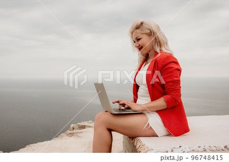 Digital nomad, a business woman in red blazer with a laptop sits on the rocks by the sea during sunset, makes a business transaction online from a distance. Freelance, remote work on vacation. Digital nomad, a business woman in red blazer with a laptop sits on the rocks by the sea during sunset, makes a business transaction online from a distance. Freelance, remote work on vacation. 96748415