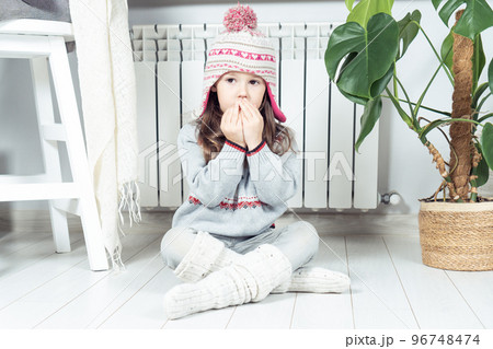 Little brunette girl breathing, inhaling on hands to keep warm near heating radiator, convector indoors. Cold apartment 96748474