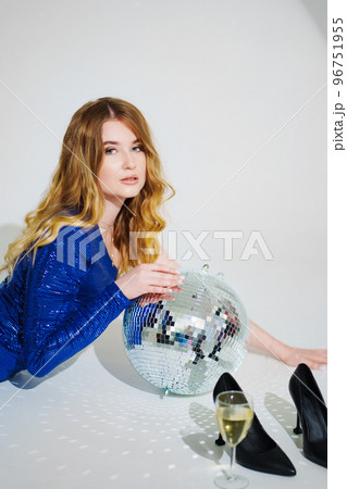 woman in shiny blue dress on floor with champagne and disco ball on a white room woman in shiny blue dress on floor with champagne and disco ball on a white room 96751955