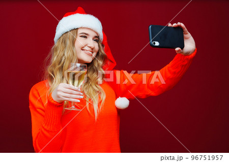 A woman in a Santa Claus hat with champagne and smartphone on a red background. 96751957