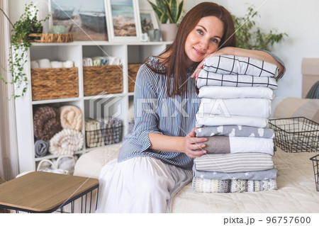 Beautiful woman housewife sits in the bedroom and stacks cotton and linen bedding and towels in a high pile Beautiful woman housewife sits in the bedroom and stacks cotton and linen bedding and towels in a high pile 96757600