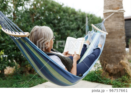 Smiling senior good-looking grey hair woman wearing glasses while reading in hammock in the summer garden 96757661
