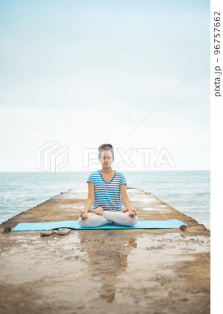 Woman yoga instructor sitting in lotus posture hands mudra admiring sea coastline landscape 96757662