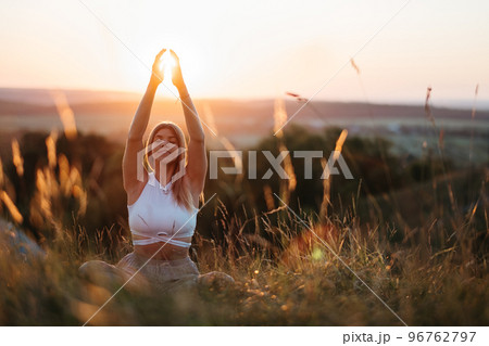 Young Woman Sitting in Meditation Yoga Pose and Catching Sun by Hands at Sunset 96762797