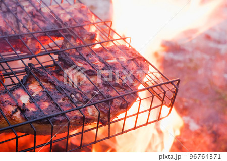 Cooking chicken meat over charcoal. Chicken meat fried with a crust on a wire rack during grill cooking. Backlight from a burning fire and smoke in the late evening. 96764371
