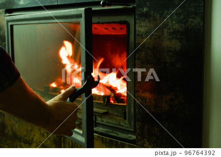 A man's hand opens the glass door of the fireplace in which the wood is burning. Modern closed fireplace with glass. A man's hand opens the glass door of the fireplace in which the wood is burning. Modern closed fireplace with glass. 96764392