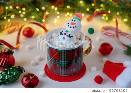 Preparing for Christmas. A red plaid mug with Snowman and marshmallows on a background of Christmas decorations and fir branches with an illuminated garland. Winter Christmas scene. 96768105