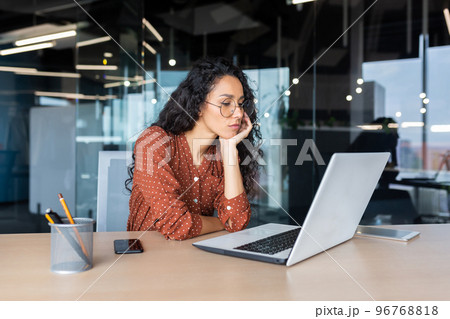 Sad grumpy Latin American woman working inside the office, business woman is bored looking at the laptop screen. 96768818