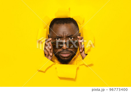 Serious african american man looking at camera through sunglasses, breaking through yellow paper background 96773400