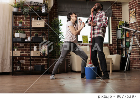 Happy multiracial couple singing with emotion, enjoying housework together. Woman singing with remote control while husband uses mop as microphone, doing spring cleaning with good vibes. Happy multiracial couple singing with emotion, enjoying housework together. Woman singing with remote control while husband uses mop as microphone, doing spring cleaning with good vibes. 96775297