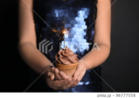 Woman holding birthday cupcake with sparkler on black background, closeup Woman holding birthday cupcake with sparkler on black background, closeup 96777558