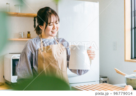 朝キッチンで電気ケトルを持ってコーヒーを淹れるエプロン姿のアジア人女性 96779014