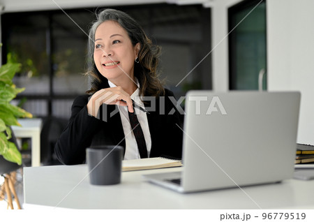 Cheerful Asian aged businesswoman daydreaming, smiling and looking out the window. Cheerful Asian aged businesswoman daydreaming, smiling and looking out the window. 96779519
