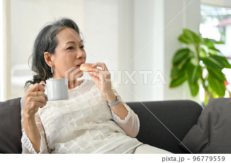 Relaxed elderly Asian woman enjoys sipping coffee an eating doughnut while chilling in living room. 96779559