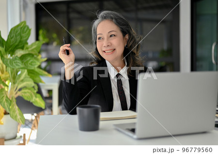 Charming Asian senior female manager sitting at her desk, smiling, daydreaming about her success. Charming Asian senior female manager sitting at her desk, smiling, daydreaming about her success. 96779560