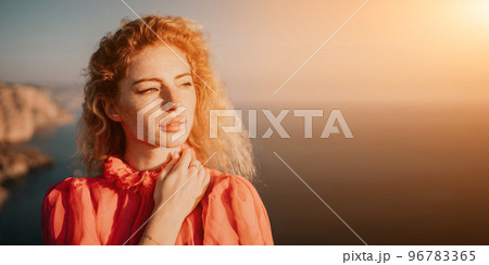 Close up portrait of curly redhead young caucasian woman with freckles looking at camera and smiling. Cute woman portrait in a pink long dress posing on a volcanic rock high above the sea at sunset Close up portrait of curly redhead young caucasian woman with freckles looking at camera and smiling. Cute woman portrait in a pink long dress posing on a volcanic rock high above the sea at sunset 96783365