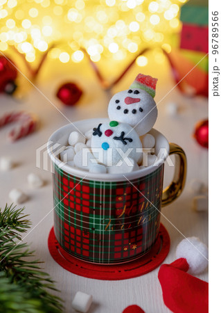 Christmas Red mug with marshmallow snowman. Preparing for Christmas. A smiling marmalade Snowman in a marshmallow mug against a backdrop of Christmas decorations and glowing garland. 96789566