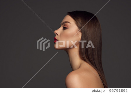 Profile view of female fashion model with naked shoulders and long hair looking at camera over dark grey background. Beautiful woman with trendy make-up, red lipstick and well-kept skin. 96791892