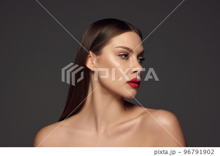 Portrait of female fashion model with naked shoulders and long hair looking away over dark grey background. Beautiful woman with trendy make-up, red lipstick and well-kept skin. Portrait of female fashion model with naked shoulders and long hair looking away over dark grey background. Beautiful woman with trendy make-up, red lipstick and well-kept skin. 96791902