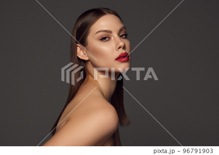 Portrait of female fashion model with naked shoulders and long hair looking at camera over dark grey background. Beautiful woman with trendy make-up, red lipstick and well-kept skin. Portrait of female fashion model with naked shoulders and long hair looking at camera over dark grey background. Beautiful woman with trendy make-up, red lipstick and well-kept skin. 96791903