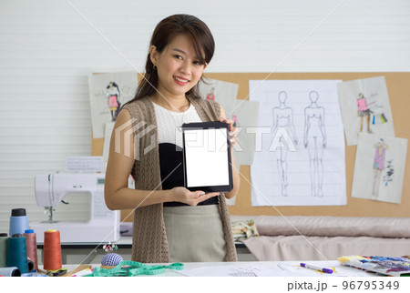 Fashion designer room. Young dressmaker with a smile showing blank screen tablet computer infront. Sewing Machine and various sewing related items are on the table. 96795349