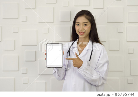 Young asian doctor in white gown and stethoscope pointing finger at white screen tablet computer, standing in front of white rectangle pattern ceramic wall. Young asian doctor in white gown and stethoscope pointing finger at white screen tablet computer, standing in front of white rectangle pattern ceramic wall. 96795744
