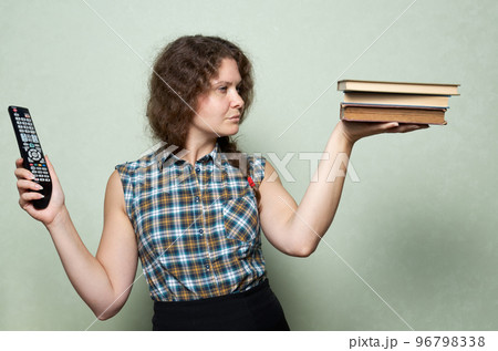 Book vs television. A woman makes a choice between books in one of her hands and a TV remote control in the other Book vs television. A woman makes a choice between books in one of her hands and a TV remote control in the other 96798338