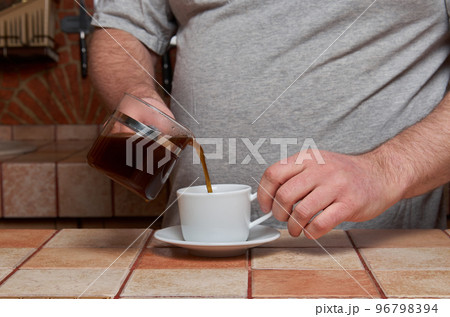 Male hands pour coffee into a coffee cup standing on a tiled kitchen table 96798394